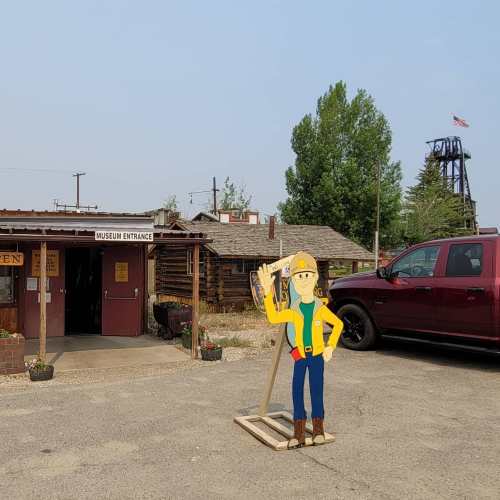 A museum entrance with a wooden sign, a cartoon construction worker cutout, and a red truck parked nearby.