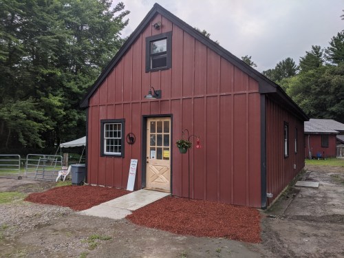 A red barn-style building with a front door, windows, and a small garden area, surrounded by trees.