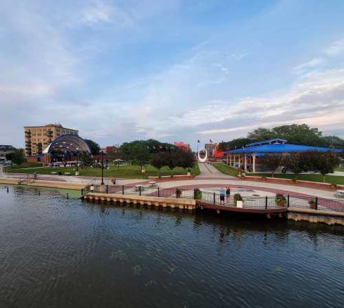 A scenic view of a park by a river, featuring green lawns, a stage, and a blue sky with scattered clouds.