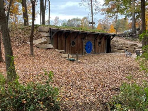 A rustic wooden structure with a blue circular door, surrounded by autumn leaves and trees in a wooded area.