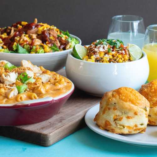 A colorful spread of dishes including salads, macaroni, and cheesy biscuits, with drinks in the background.