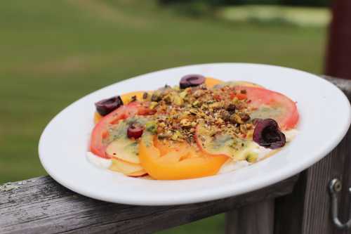 A plate of colorful sliced tomatoes topped with nuts and herbs, set against a green outdoor background.