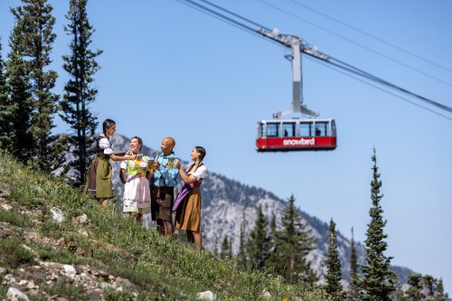 A group of four people in traditional attire enjoy drinks outdoors, with a cable car passing overhead in a mountainous area.