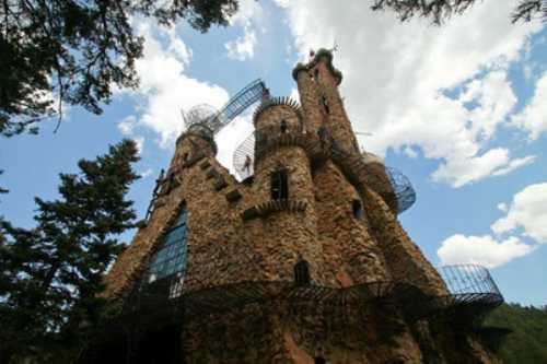 A whimsical stone castle with towers and metal balconies, surrounded by trees under a partly cloudy sky.