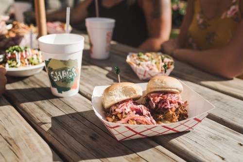 Two pulled pork sandwiches with coleslaw on a wooden table, accompanied by a drink and people in the background.