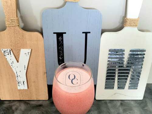 A pink smoothie in a glass sits between three decorative wooden boards with the letters Y, U, and M.