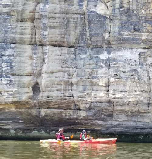 Two people in a kayak paddle along a river beside a large, rocky cliff.