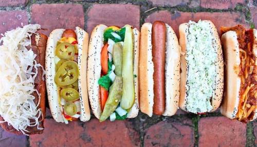A colorful array of hot dogs with various toppings, including sauerkraut, pickles, and vegetables, on a brick surface.