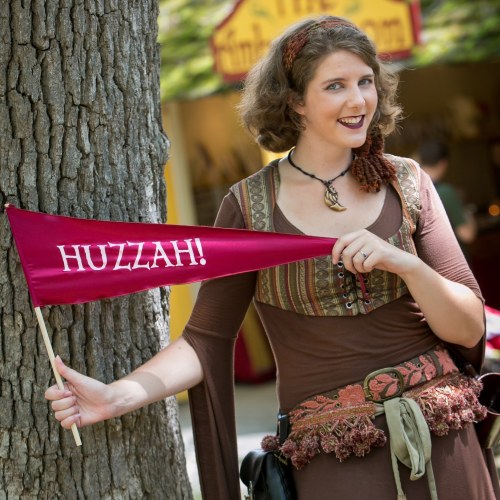 A woman in a medieval-style outfit holds a flag that says "HUZZAH!" while standing by a tree.