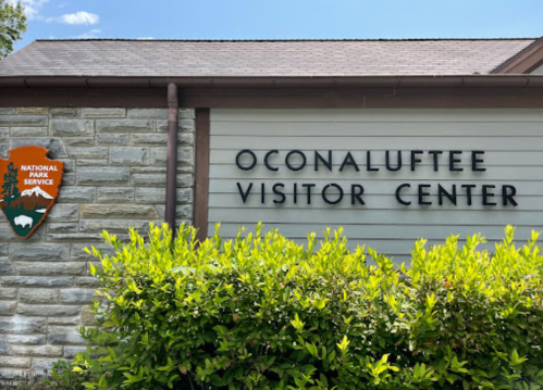 Sign for Oconaluftee Visitor Center with National Park Service logo, surrounded by greenery and a clear blue sky.