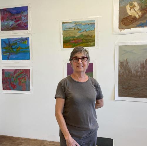 A woman with short gray hair stands in an art studio, surrounded by colorful paintings on the walls.