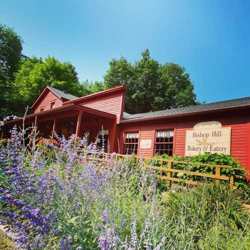 A red building with a sign reading "Bishop Hill Bakery & Eatery," surrounded by greenery and colorful flowers.