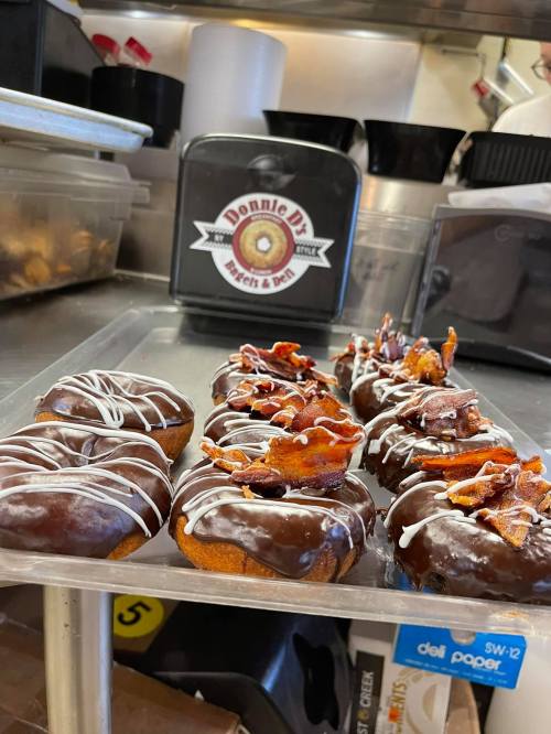 A tray of chocolate-covered donuts topped with bacon and drizzled with white icing, displayed in a kitchen setting.