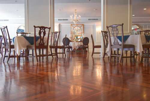 Elegant dining room with wooden floors, tables set with white linens, and a chandelier in the background.