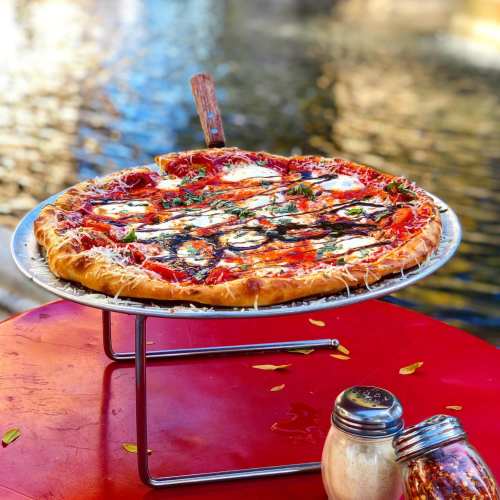 A freshly baked pizza with cheese and basil on a red table, beside a salt shaker, near a body of water.