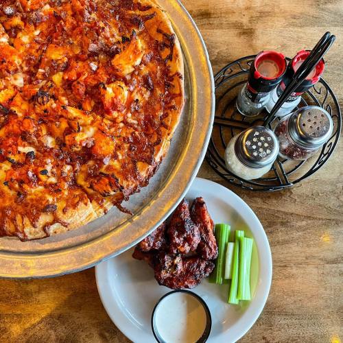 A pizza topped with cheese and toppings, served with chicken wings, celery sticks, and ranch dressing on a wooden table.