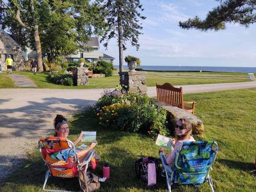 Two women sit in colorful chairs, painting by a garden near the shore, with a scenic view of the ocean in the background.