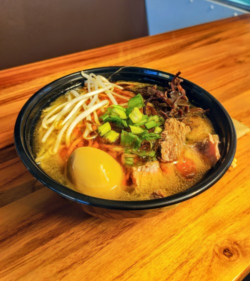 A bowl of ramen with broth, sliced meat, green onions, bean sprouts, and a soft-boiled egg on a wooden table.