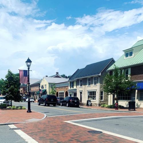 A quaint town square with brick pavement, shops, and a flagpole under a blue sky with scattered clouds.