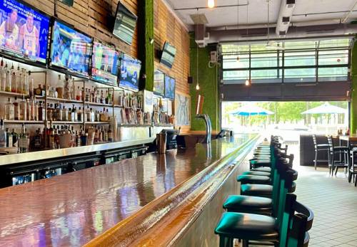 A modern bar with a polished wooden counter, multiple TVs, and a row of green chairs, overlooking an outdoor seating area.