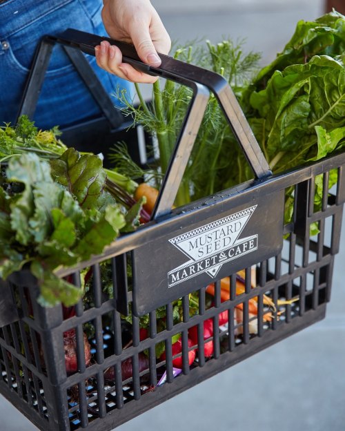 A person holds a black basket filled with fresh vegetables and greens from Mustard Seed Market & Café.