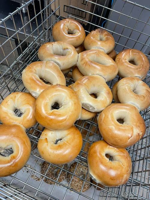 A metal basket filled with freshly baked bagels, some with a golden-brown crust, resting on a surface with scattered seeds.