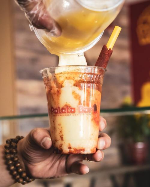 A hand pours a yellow drink into a clear cup filled with creamy dessert and a tamarind stick, showcasing vibrant colors.