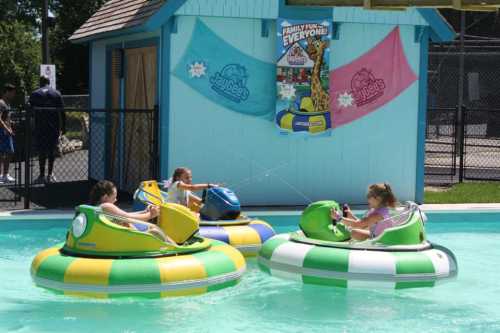 Four children in colorful bumper boats play in a pool, surrounded by a vibrant water park backdrop.