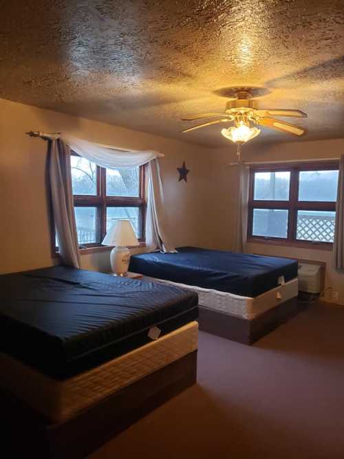 A cozy bedroom with two unmade beds, a ceiling fan, and large windows letting in natural light.