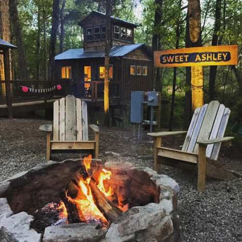 A cozy campfire in front of a wooden cabin surrounded by trees, with a sign reading "Sweet Ashley."