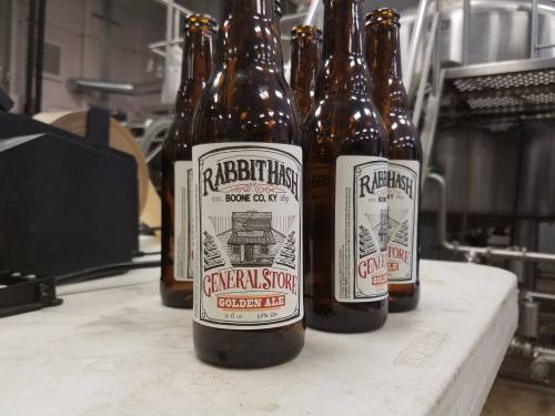 Bottles of Rabbit Hash General Store Golden Ale on a table, with brewing equipment in the background.