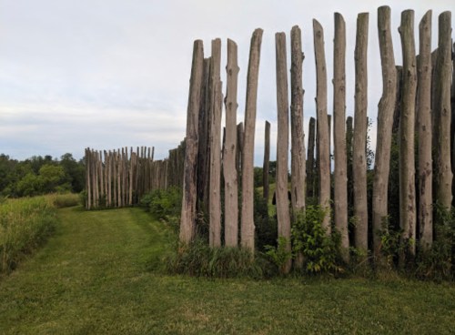 A pathway lined with tall wooden posts, surrounded by lush greenery and a cloudy sky.