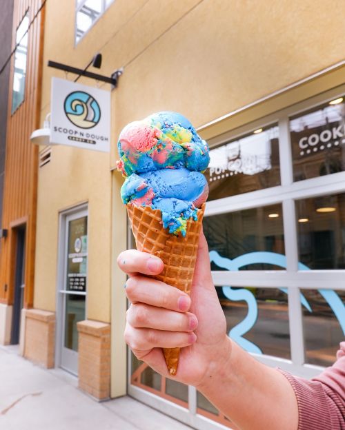 A hand holds a colorful ice cream cone with blue and rainbow swirls outside a shop named Scoopndough.