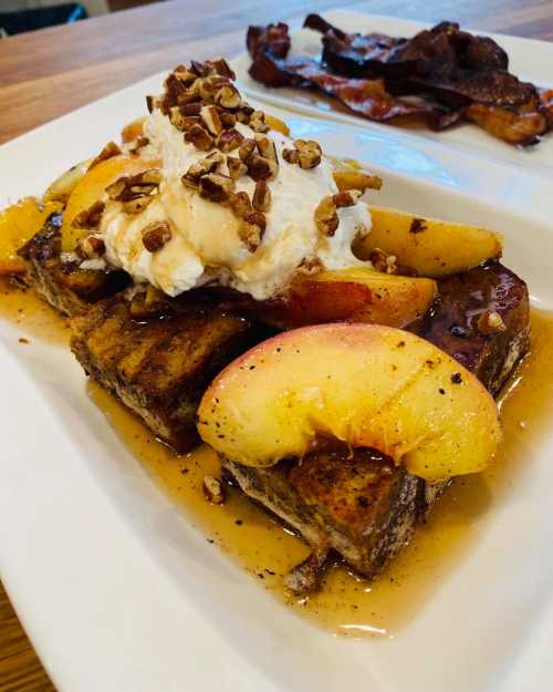 Delicious French toast topped with sliced peaches, whipped cream, pecans, and drizzled with syrup, served with bacon.