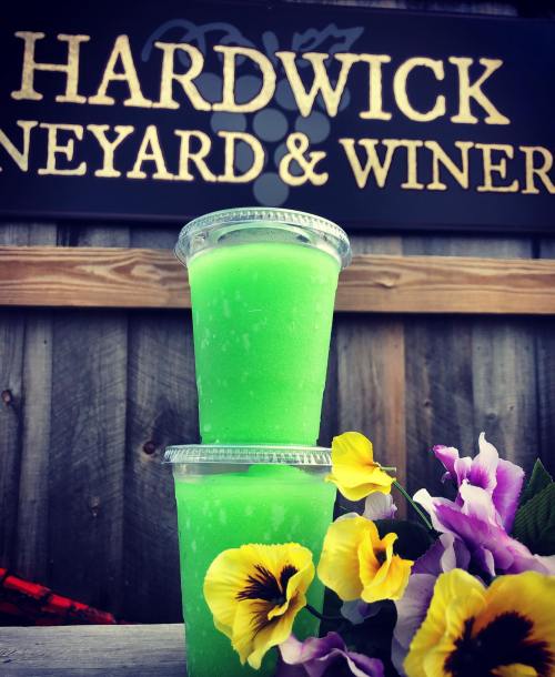 Two green drinks in clear cups sit in front of a sign for Hardwick Vineyard & Winery, with colorful flowers nearby.