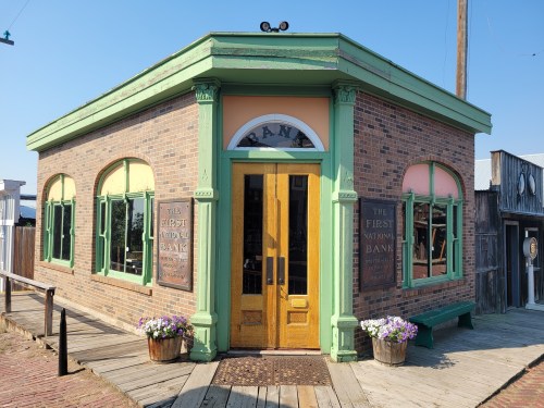Historic bank building with green trim, wooden doors, and flower pots, set in a rustic outdoor environment.