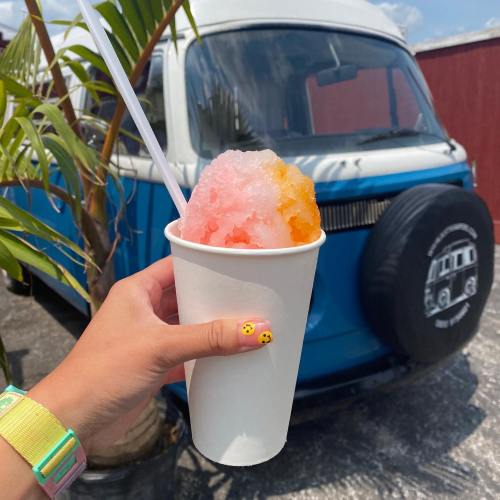A hand holds a cup of colorful shaved ice in front of a blue vintage van and palm tree.