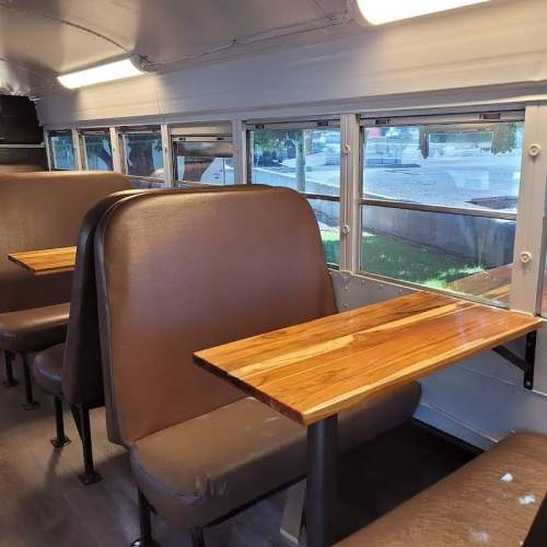 Interior of a bus with brown leather seats and wooden tables, featuring large windows for natural light.