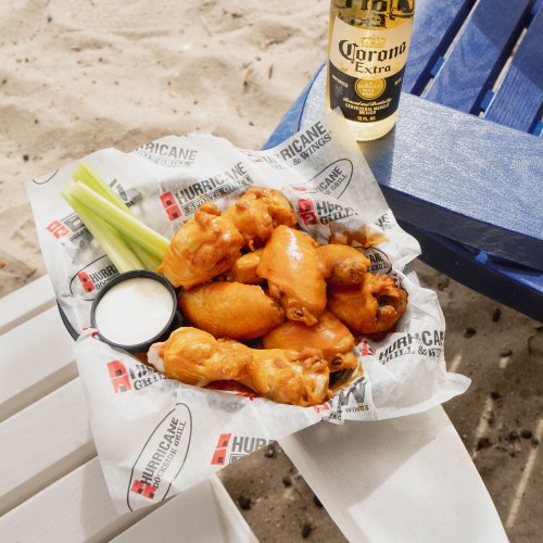 A plate of chicken wings with celery and ranch dressing, alongside a bottle of Corona beer on a sandy beach.