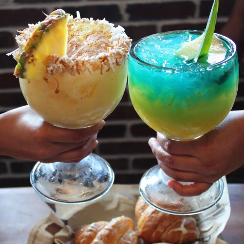 Two colorful cocktails in large glasses, one with a pineapple garnish, held by hands against a brick background.