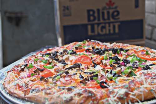 A close-up of a delicious pizza topped with tomatoes, olives, green peppers, and cheese, with a beer box in the background.