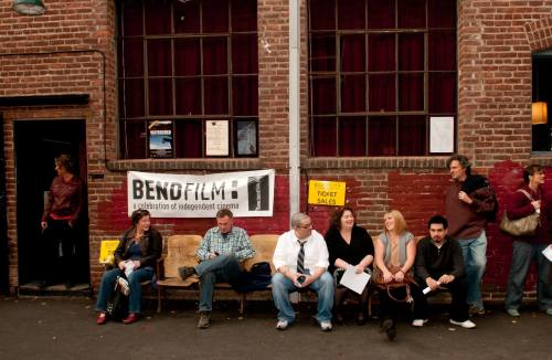 A group of people sitting on a bench outside a building, with a sign for BendFilm and ticket sales visible.