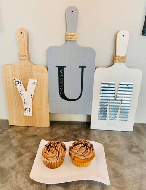 Three decorative cutting boards spell "YUM" above two chocolate frosted cupcakes on a white plate.