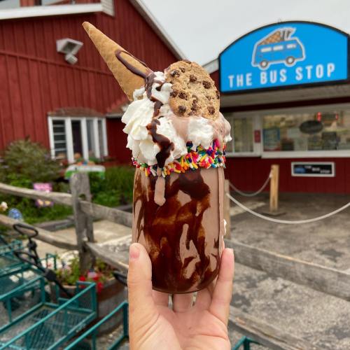 A hand holds a tall glass filled with ice cream, chocolate syrup, whipped cream, a cookie, and a cone, in front of a shop.
