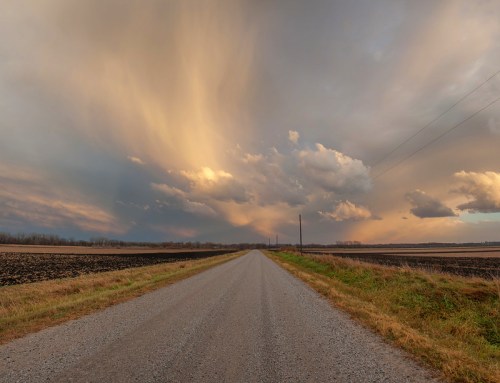 A gravel road stretches into the distance under a dramatic sky with colorful clouds at sunset. Fields line the sides.