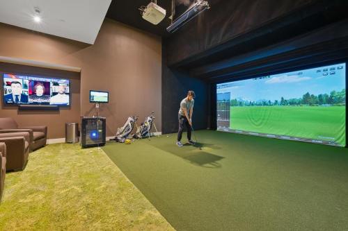 A person practicing golf in a home simulator room with a large screen, green turf, and a TV displaying news.