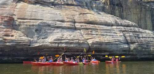 A group of kayakers in red and yellow kayaks paddling near a rocky cliff on a calm waterway.