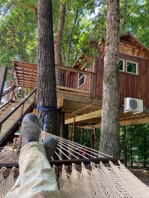 A person relaxes in a hammock under a treehouse, surrounded by trees and nature.