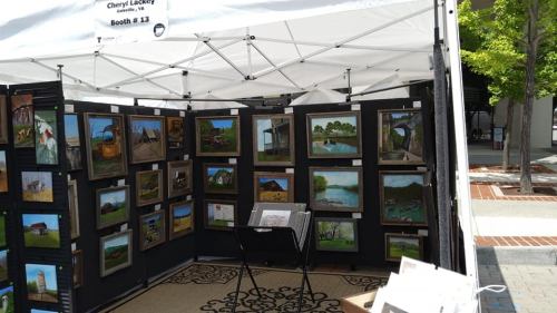 Art booth featuring framed paintings displayed on black panels under a white tent, with a chair and decorative rug.
