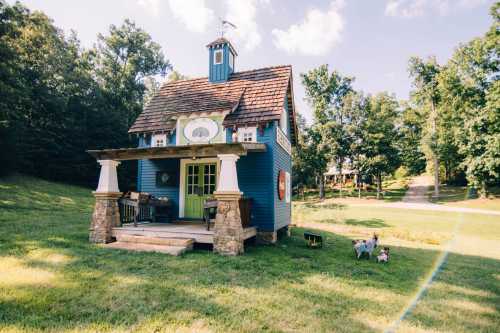 A charming blue cottage with a porch, surrounded by greenery and two small dogs in the foreground.